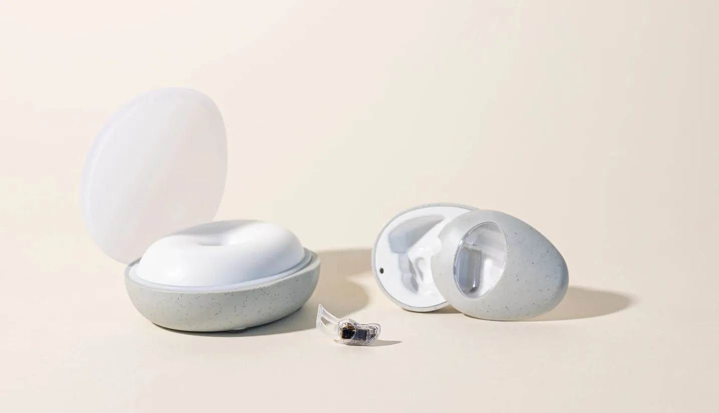 Studio photo of the speckled grey pebble base station with its frosted lid open, a second unit tipped on its side, and the wearable between them on a cream background