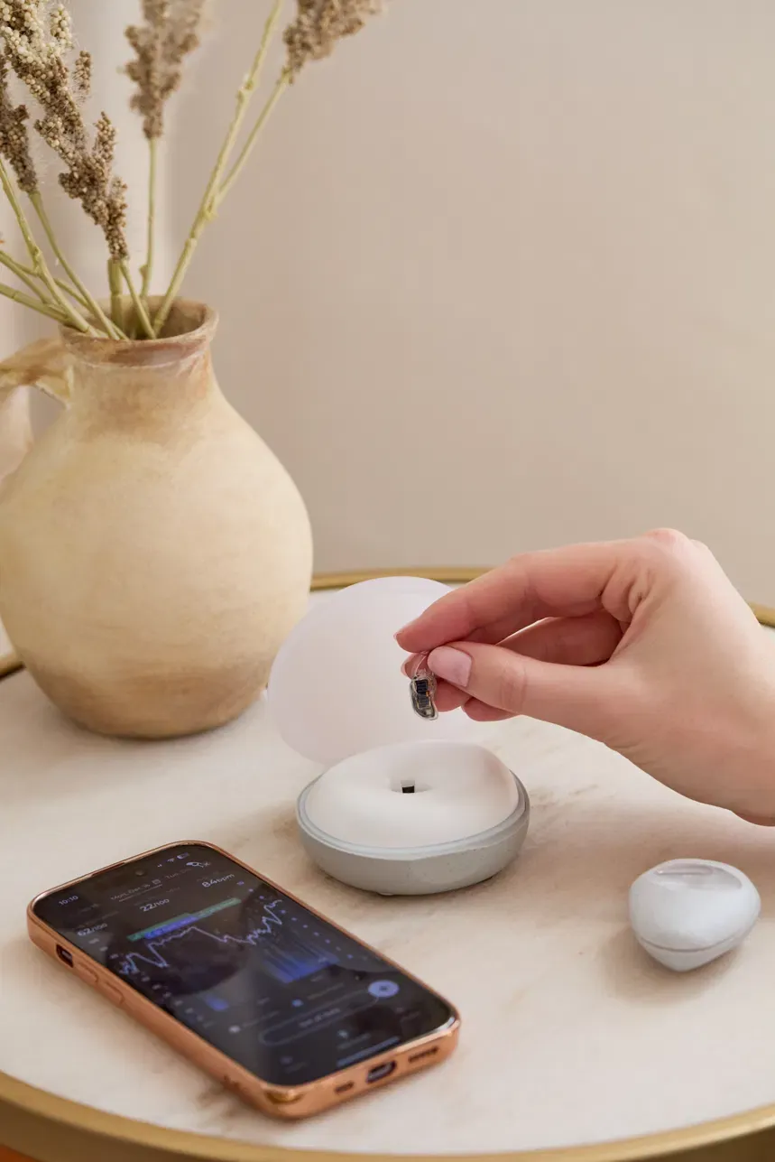 A hand lifting the wearable from the base station on a small round table, next to a phone showing the Lumia app