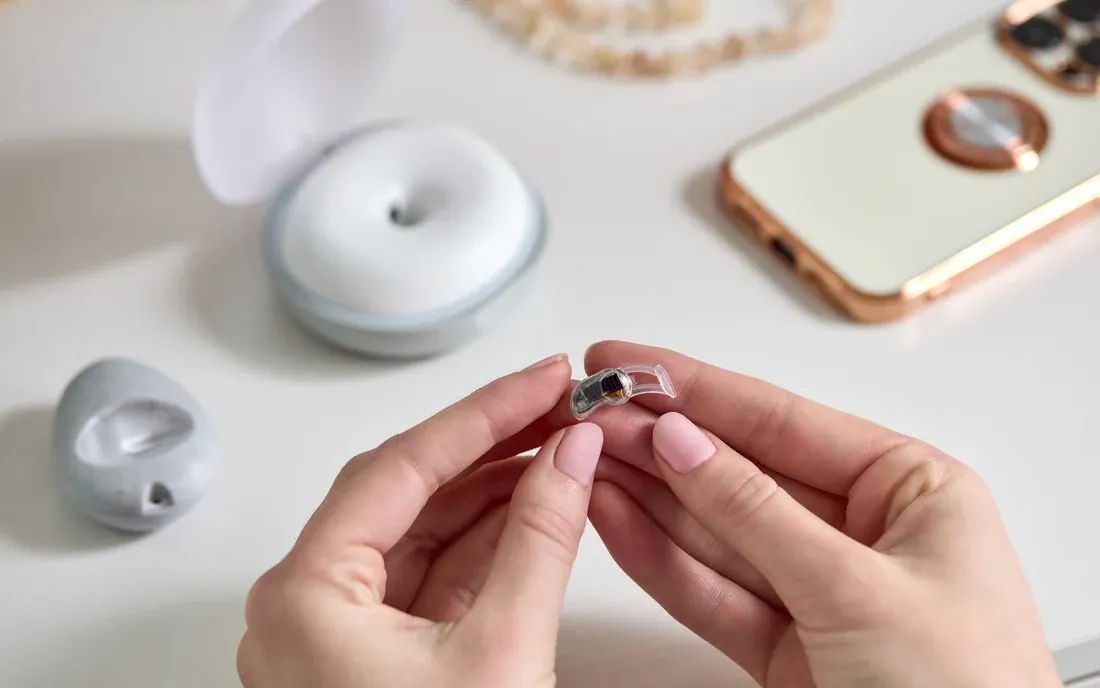 Two hands holding the small clear in-ear wearable over a white table, with the open pebble-shaped base station and a phone behind