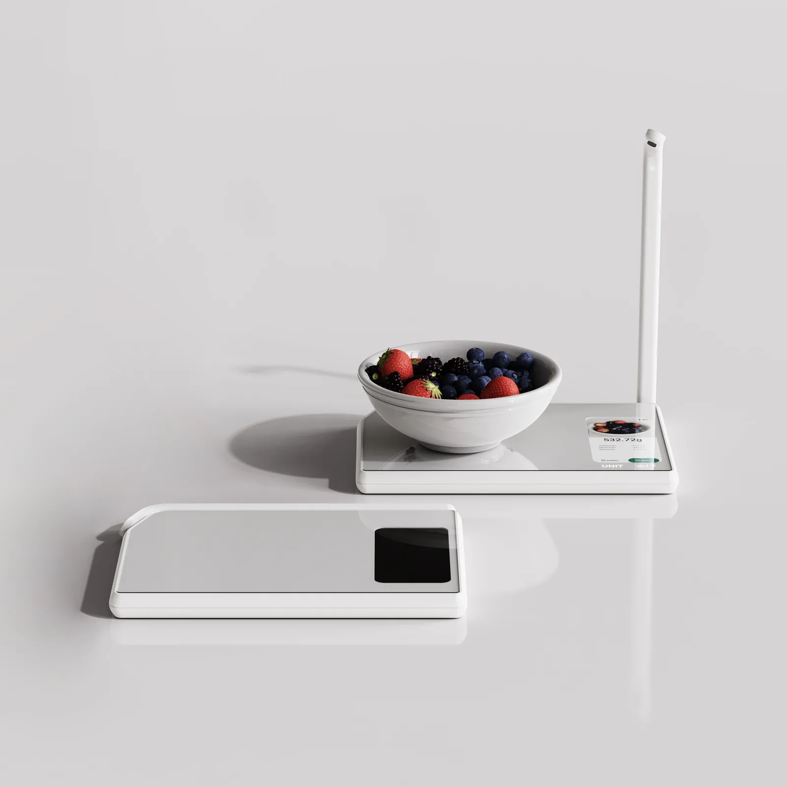 Two white Nutri scales on a grey counter: one open with the camera arm raised and a bowl of berries on its glass top, one folded flat in front with the arm lying across the base