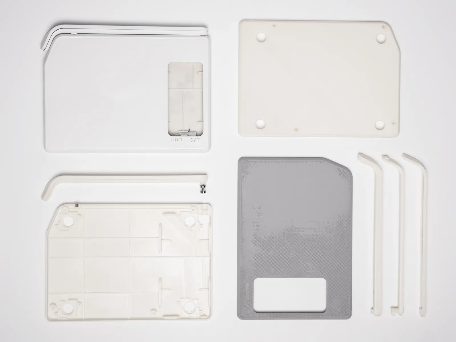 Flat lay of 3D printed scale parts on a white ground: the top shell with its screen cutout and UNIT and power/tare marks, the bottom shell with ribs and screw bosses, a printed camera arm with its pin, a flat base plate, a grey plate with a window for the load cell, and three arm sections