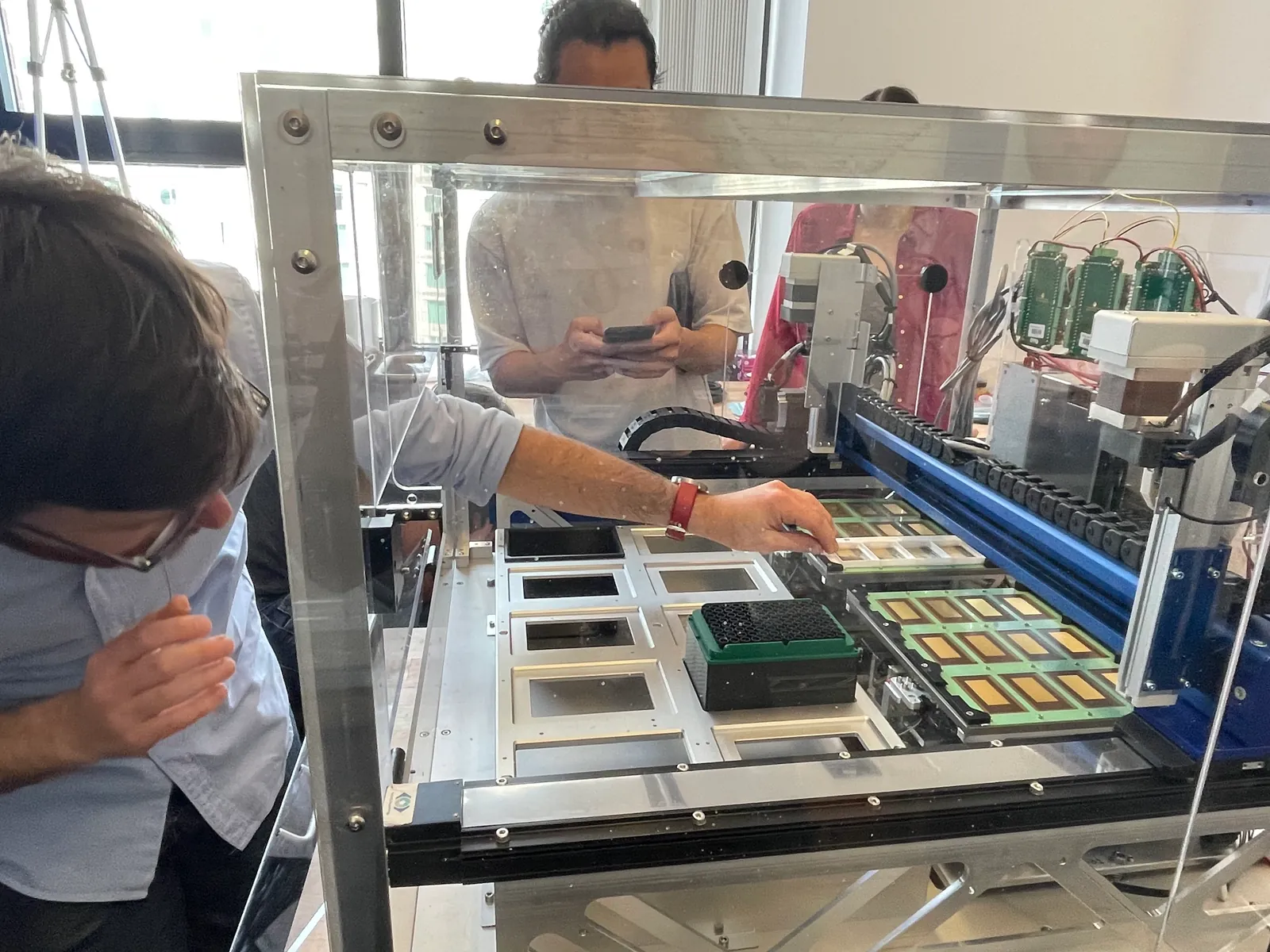 The initial machine in an acrylic enclosure on a bench, with the deck of labware trays and the gantry visible, and engineers working around it