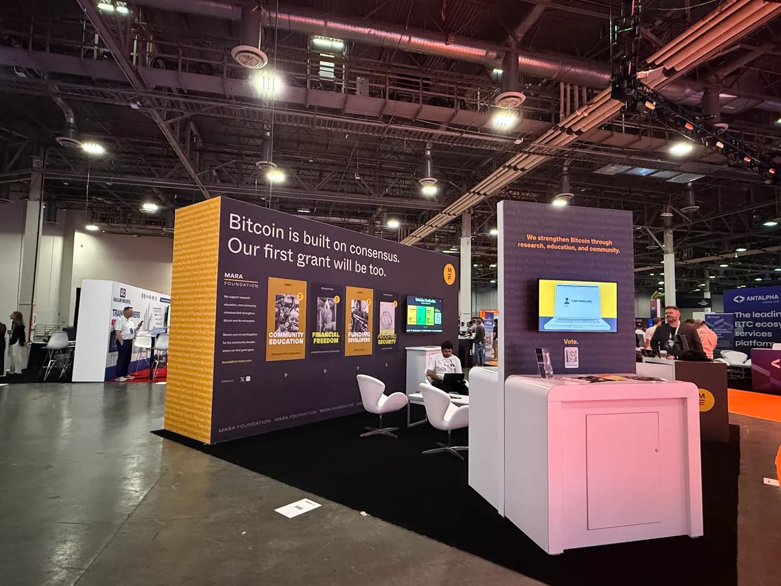 Booth wall reading Bitcoin is built on consensus, our first grant will be too, with three poster panels, chairs, and a voting kiosk