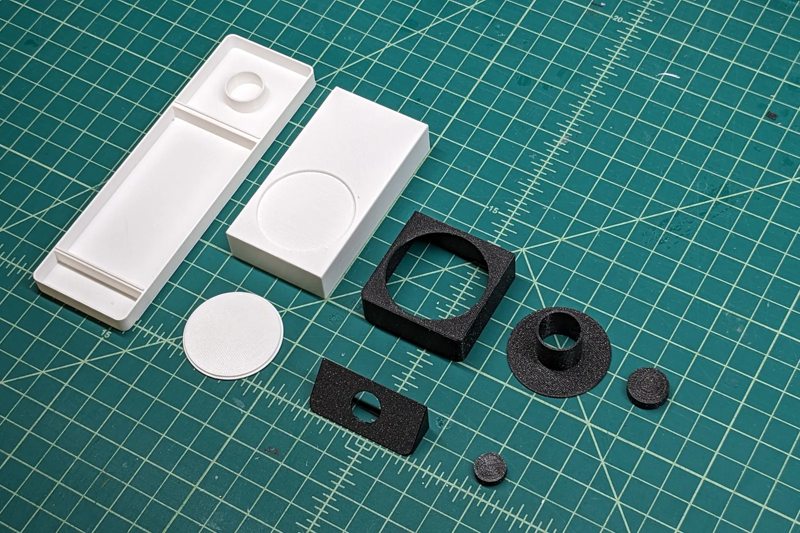 3D printed parts laid out on a cutting mat: white body shell, camera block, button disc, and black chin, ring and trim pieces